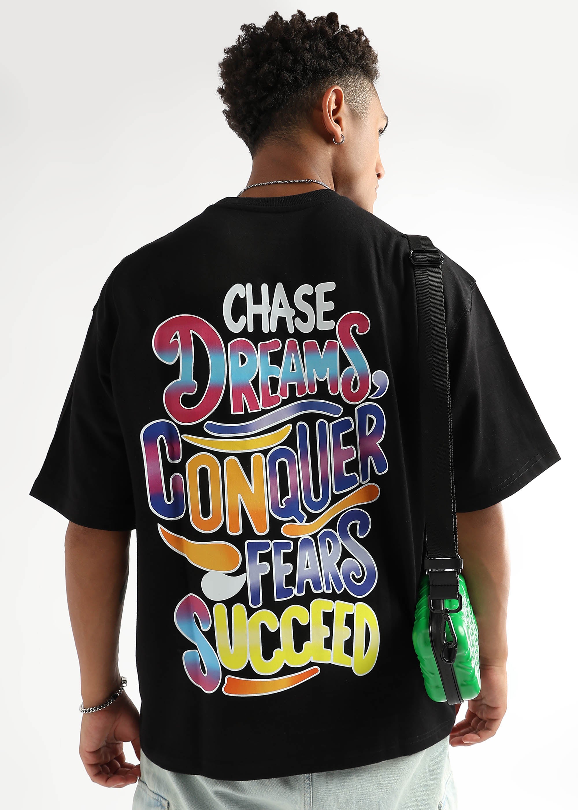 A person wearing an oversized black t-shirt with a bold graphic on the back that reads 'CHASE DREAMS, CONQUER FEARS, SUCCEED' in multicolored letters.